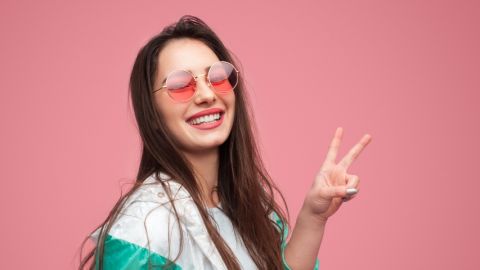 Une jeune femme sur fond rose en lunettes rose fait un signe peace des doigts