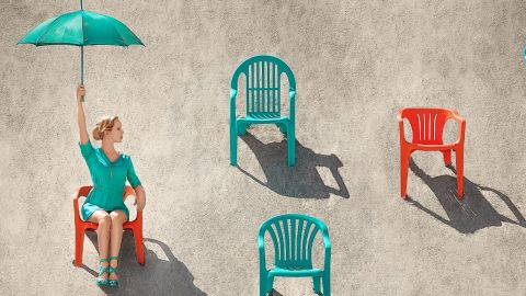 Jeune femme blonde tenant un parasol turquoise sur une plage, assise à travers de nombreuses chaises rouges et sarcelles dans le sable, par une journée ensoleillée