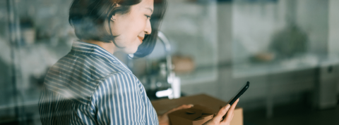 Femme asiatique souriante se tenant derrière une fenêtre, tenant son téléphone et gérant un système de notation depuis sa cuisine.