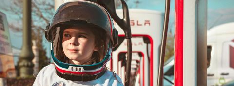 Un jeune enfant avec un casque et une tenue spatiale devant des bornes électriques Tesla