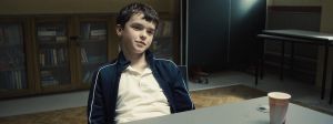 une jeune garçon assi. à une table l'air sur de lui.